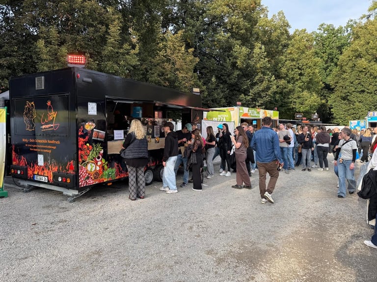 People line up at a black food truck at an outdoor event surrounded by trees