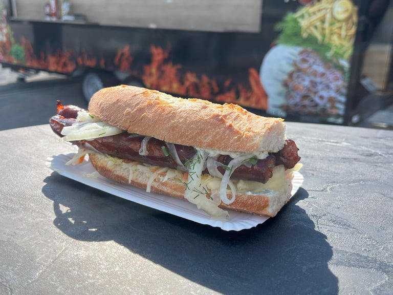 A grilled sandwich on crusty bread filled with sliced meat, cheese, and vegetables, served on a white paper wrapper at a food truck with fire graphic visible in background.