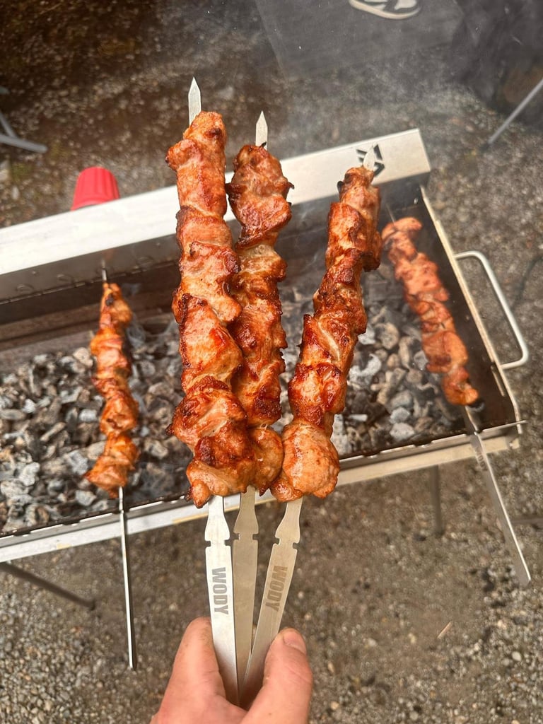 Hand holding grilled meat skewers over a portable BBQ grill with charcoal fire