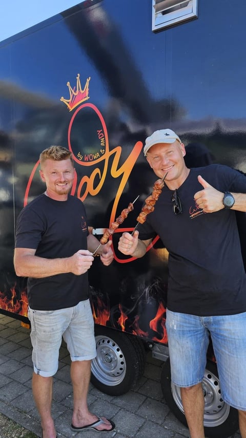 Two men holding grilled meat skewers pose in front of a food truck with flame graphics and a crown logo