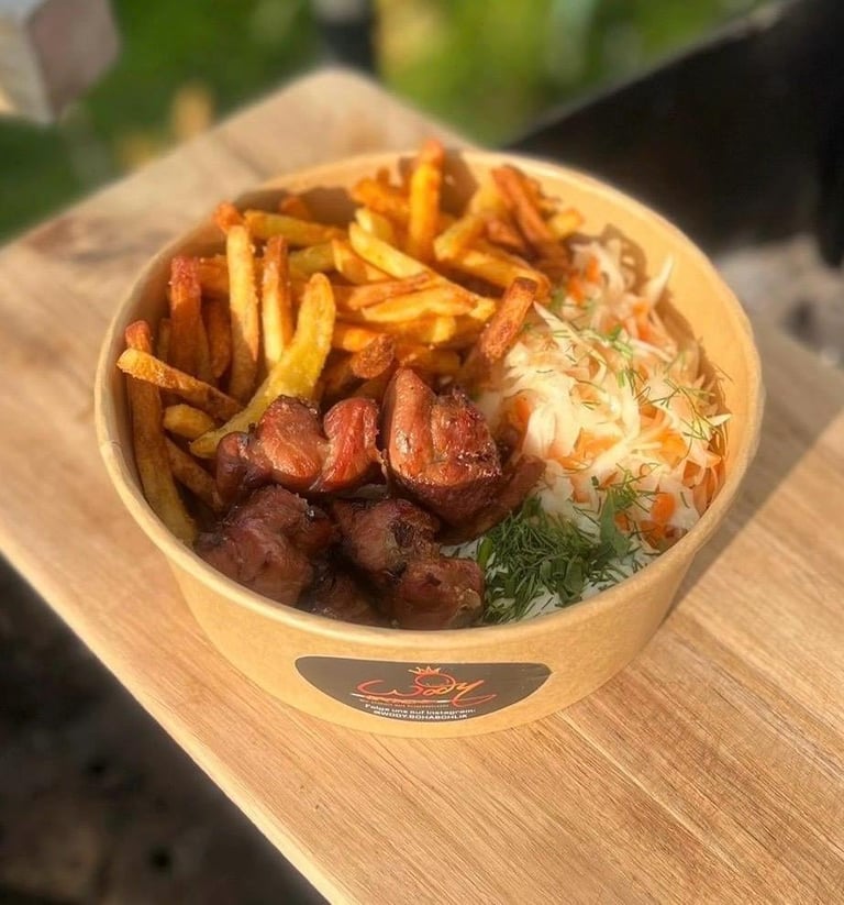 Wooden bowl containing golden fries, sliced red sausage, coleslaw, and fresh dill garnish on an outdoor table