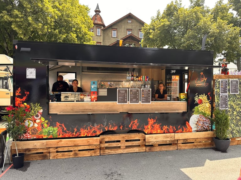 Black food truck with wooden counter and bar, red flame graphics, staff inside, potted plants and trees in background