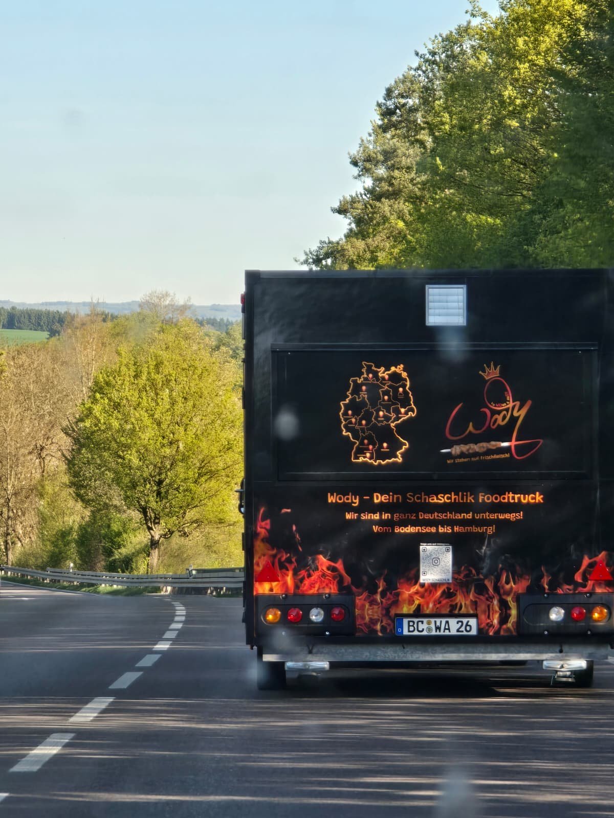 Black truck on highway with German text and flame graphics on rear, trees and landscape visible in background