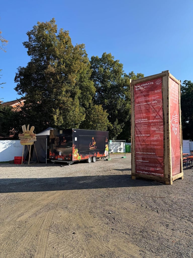 Outdoor market or festival area with food trucks and vendor booths beneath tall trees on a gravel lot under clear blue sky